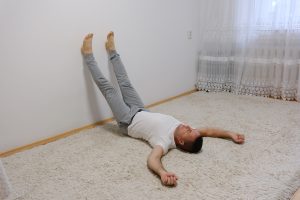 A grown man lies on the floor on a carpet at home with his legs raised up on the wall and his arms spread out to the sides.